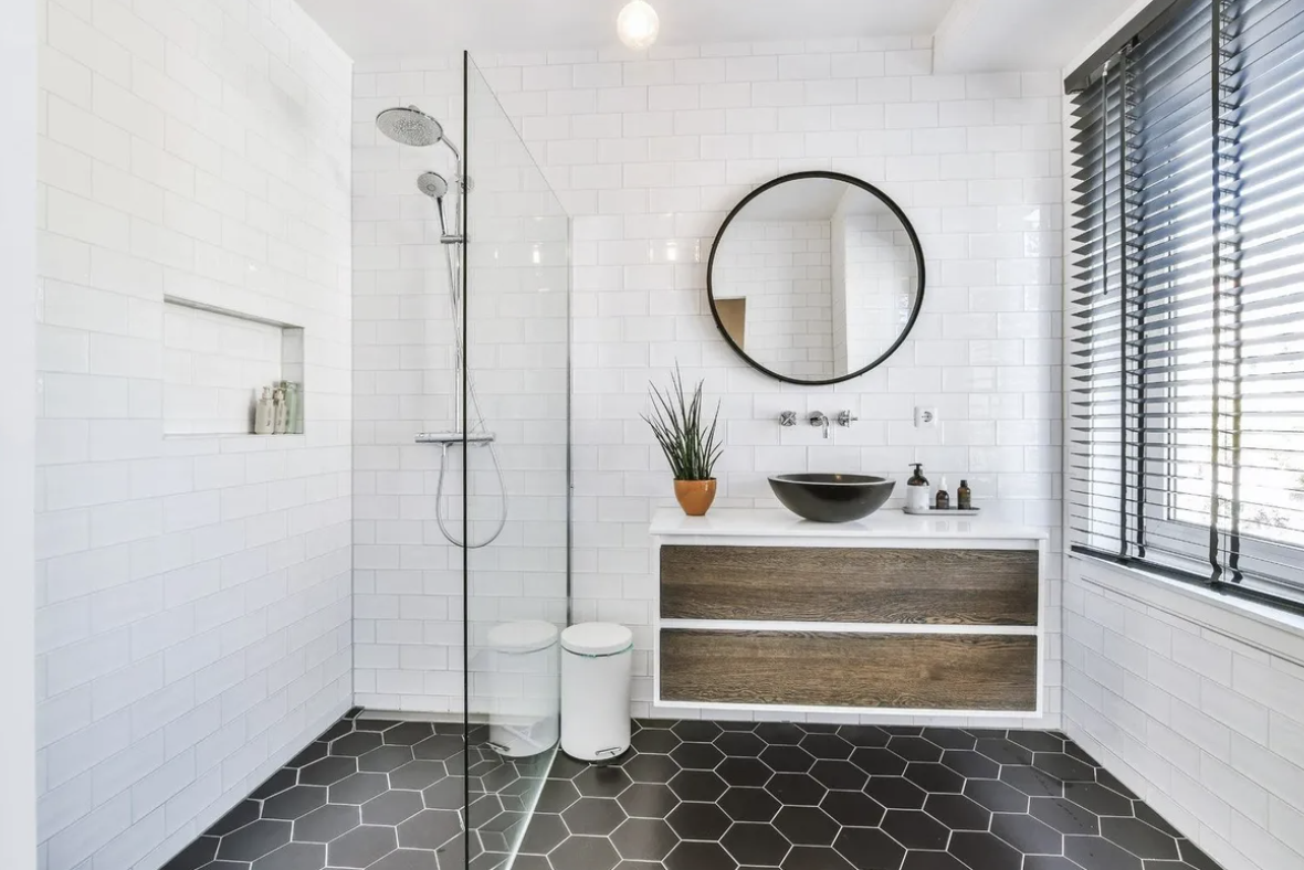 Modern bathroom with white subway tile walls, black hexagonal floor tiles, floating wood vanity, black round mirror, and glass shower enclosure