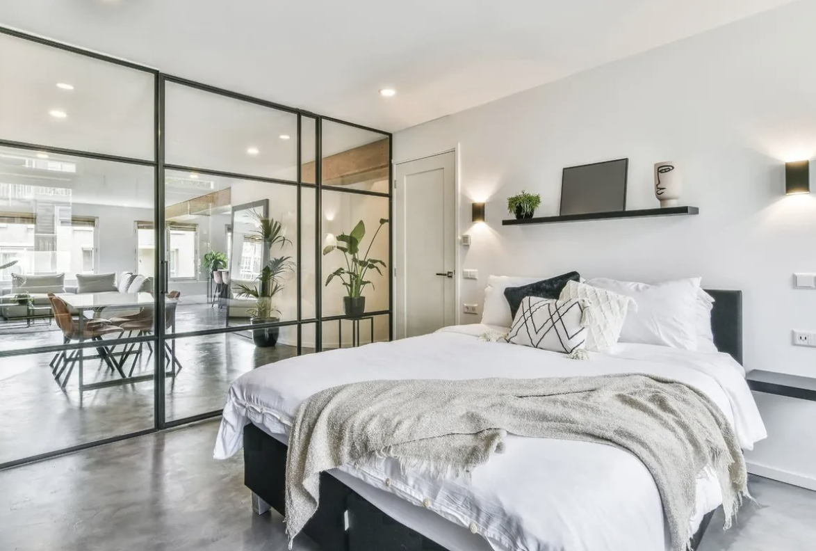 Modern bedroom with black bed, white bedding, glass partition wall separating living area, potted plants, and sleek furnishings